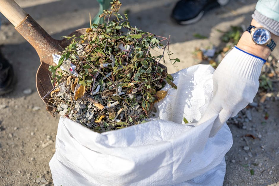 Close-up of a person wearing a white gardening glove and a wristwatch, using a metal shovel to scoop a mixture of garden debris including intertwined ivy, small branches, and dry leaves into a large w